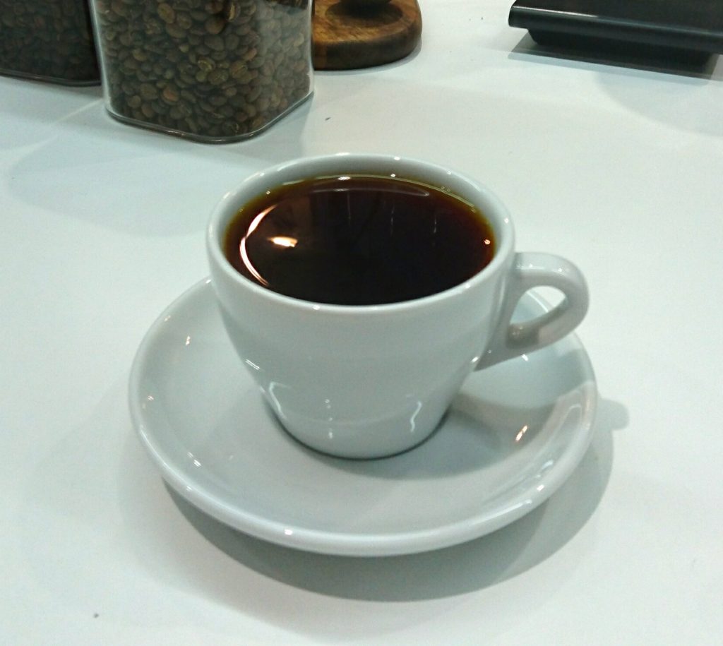 コーヒーカップに入ったコーヒーとその奥に写るコーヒー豆
